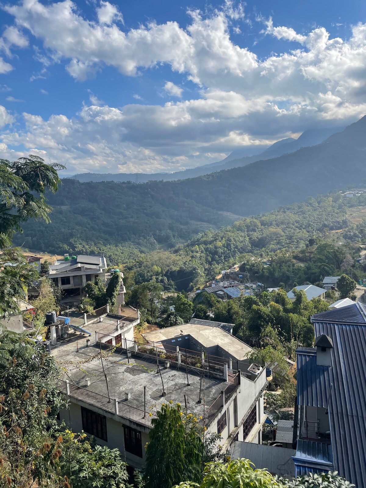 A panoramic view of the surrounding hills and valleys is depicted, showcasing a blend of greenery and rooftops. The sky is partly cloudy, allowing natural light to illuminate the landscape below. Natural terrain and scattered buildings create a serene atmosphere.