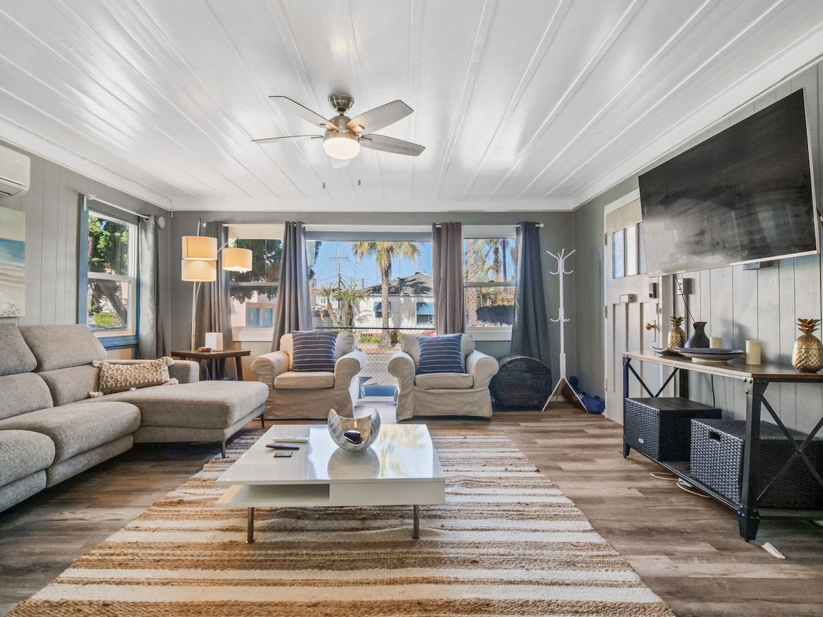 A spacious living room is filled with natural light from large windows, showcasing comfortable seating arrangements. A soft area rug covers the floor beneath a central coffee table, while a ceiling fan provides a cool breeze. Decorative elements add to the inviting ambiance.