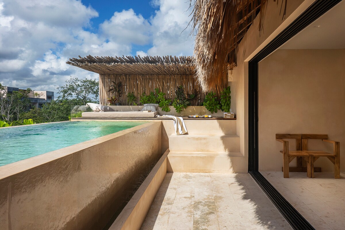 A serene private terrace features an infinity pool with a smooth edge, seamlessly blending into the lush surroundings. Natural materials are used throughout, with a thatched roof providing shade over a seating area adorned with greenery, creating a tranquil outdoor retreat.