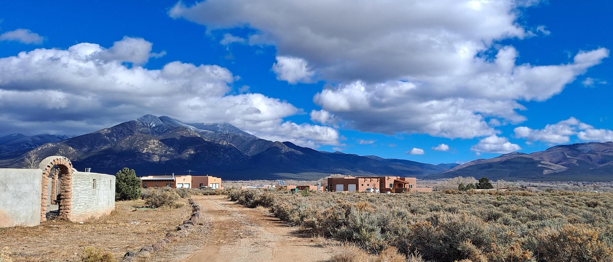A scenic view captures a wide expanse of land, featuring a dirt road leading toward adobe structures. The backdrop showcases dramatic mountains under a partly cloudy sky, with vibrant blue and white hues complementing the serene landscape.