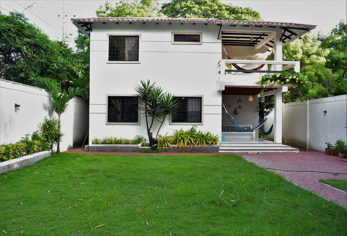 The house features a well-maintained grassy lawn in front, surrounded by tropical plants. A spacious balcony is visible on the upper level, adorned with colorful hammocks. The exterior white walls are complemented by dark wooden accents and a paved pathway leading to the entrance.