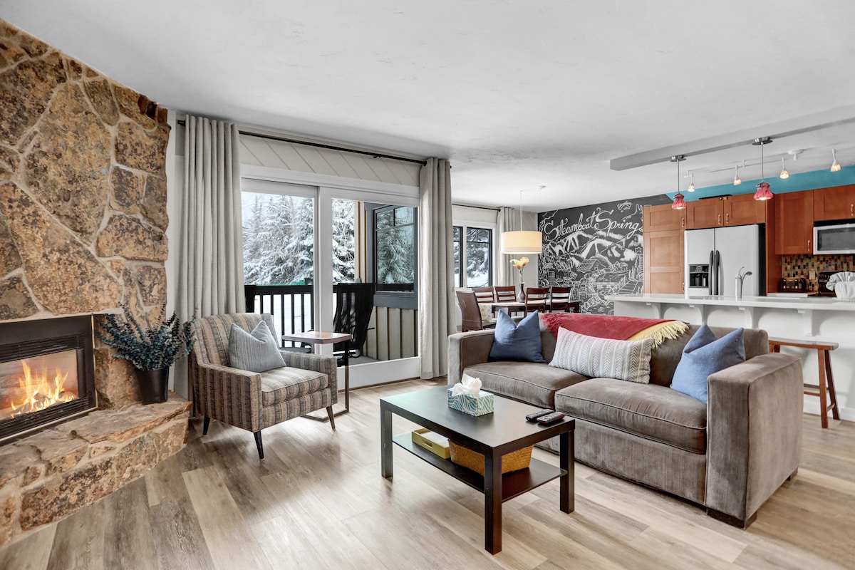 A welcoming living area is highlighted by a stone fireplace and a large window overlooking snow-covered trees. Comfortable seating includes a sofa and an accent chair, complemented by a coffee table. An open kitchen and dining area are visible in the background, featuring a sleek countertop.