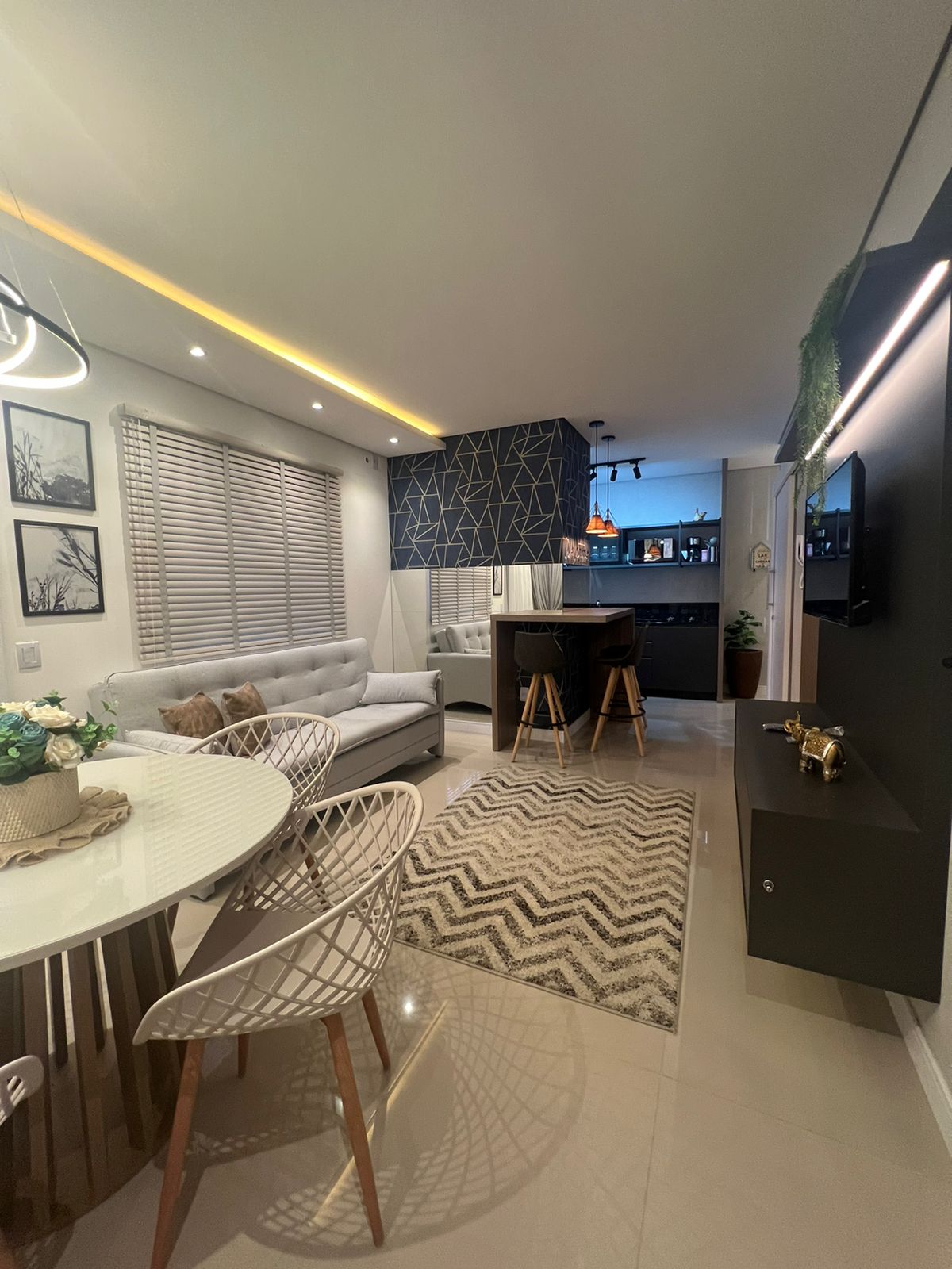 A modern living area features a plush sectional sofa and a round dining table set for six. Decorative wall art is displayed beside windows adorned with shades, while a rug adds texture to the sleek flooring. A bar counter with high stools is visible in the background.