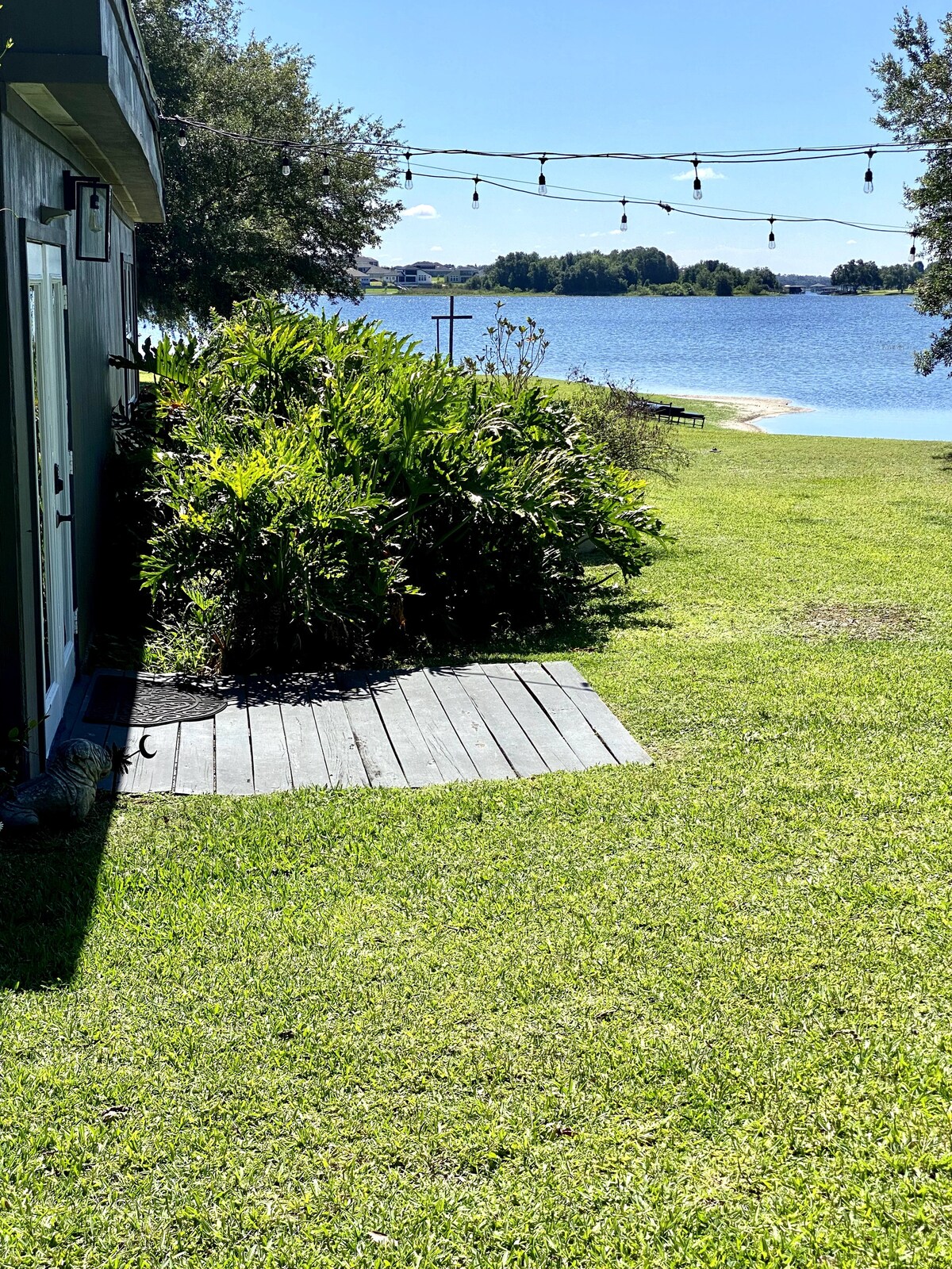 A well-maintained lawn extends toward the lake, with native greenery lining a wooden path. Strings of outdoor lights add a casual touch. The serene water is visible in the background, framed by trees, creating a tranquil outdoor atmosphere.