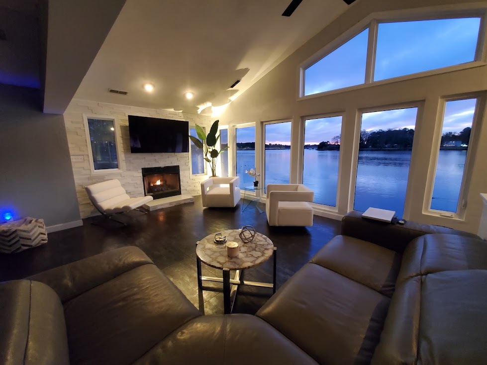 A spacious living area features large windows offering expansive views of the water. A cozy fireplace is set against a stone wall, while modern white chairs and a dark sectional provide ample seating. A coffee table sits at the center, adding to the inviting layout.