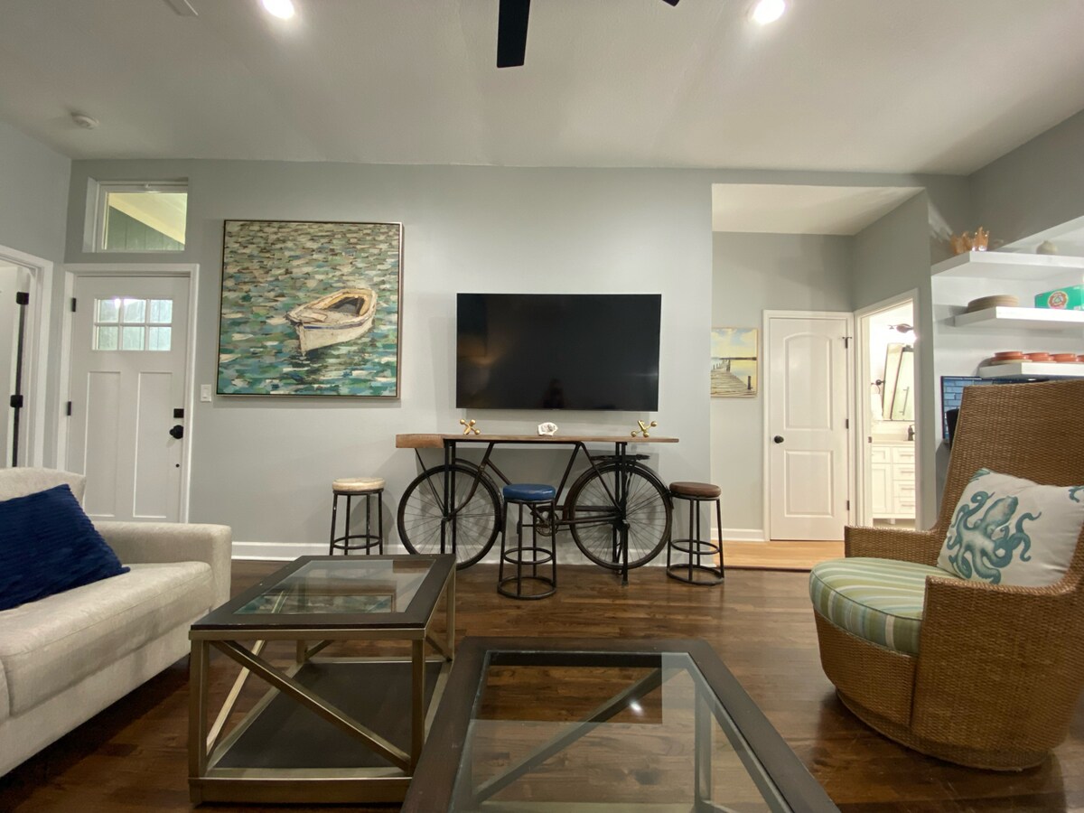 A modern living area features a large wall-mounted TV above a unique console made from bicycle wheels. A light-colored sofa, glass coffee tables, and a comfy armchair with pattern accents create a welcoming space. Natural light enters through the front door and windows.