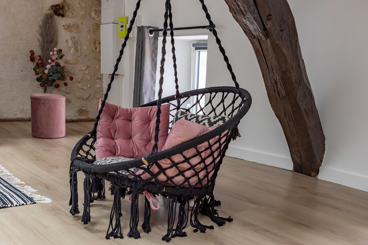 A hanging chair with a woven design is positioned in the room, featuring soft pink cushions. Natural wood beams above add character to the loft's design. The warm-toned flooring complements the space, and light enters through a nearby window, creating an inviting scene.