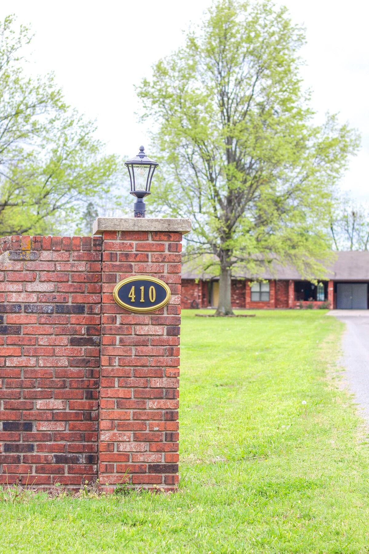 A brick column is featured at the entrance of the property, displaying the number 410. A lantern is mounted on top, providing a welcoming touch. Lush green grass surrounds the area, with a large tree visible in the background and the ranch-style home partially seen.