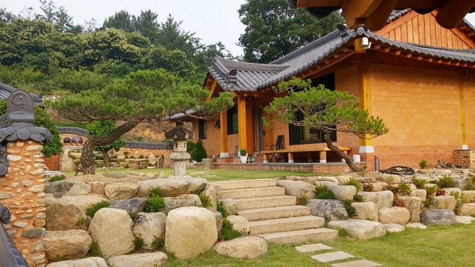 A traditional Korean house is framed by a landscaped garden featuring stone paths and carefully arranged rocks. Lush greenery, including small trees, adds tranquility to the space, while natural materials highlight the structure's earthy tones.