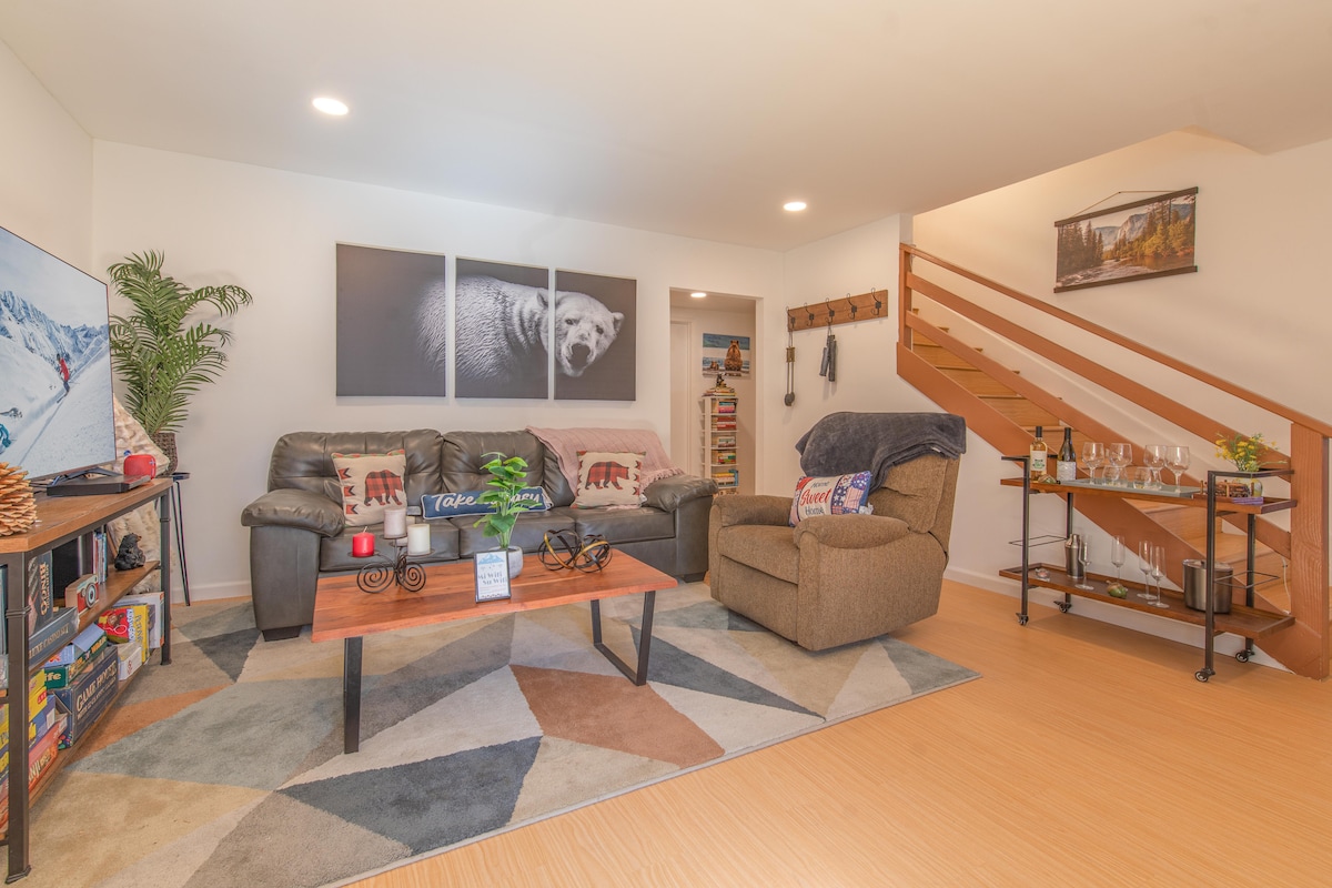 A cozy living room features a comfortable sofa and an armchair set on a geometric-patterned rug. Wall art depicting a bear is mounted above, while a television and bookshelves are visible. A staircase in the background leads to the upper level.