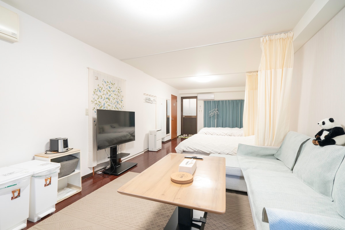 A spacious living area features a light blue sofa and wooden coffee table, with a television positioned against the wall. Two inviting beds are set in the back, complemented by curtains that add a touch of privacy. Soft lighting creates a comfortable environment.
