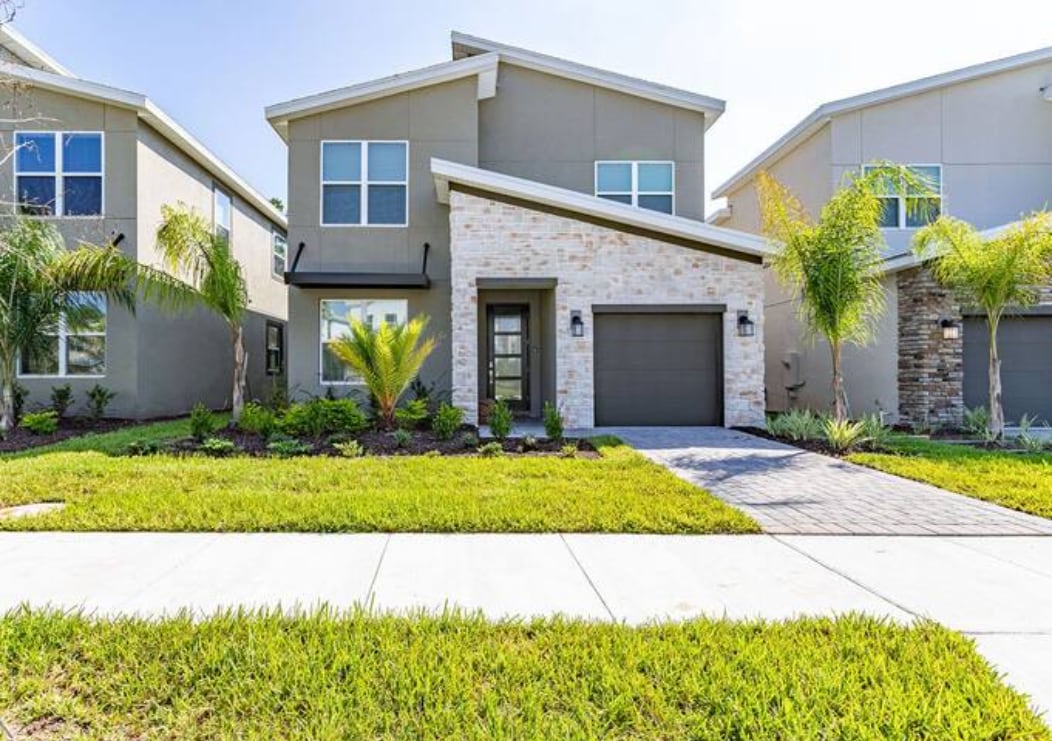 The exterior of a modern house is shown, featuring a light grey facade with stone accents. A charming pathway leads to the entrance, bordered by well-maintained landscaping that includes palm trees and shrubs. A garage door is visible on the left side of the house.