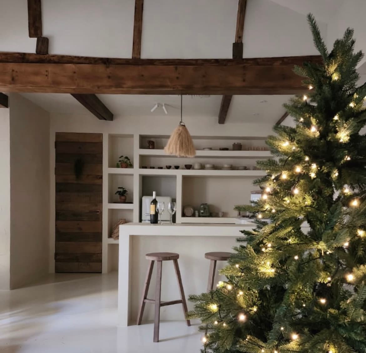 A minimalistic kitchen area is highlighted with wooden beams overhead. A tall tree adorned with warm lights stands in the foreground, while a light-colored bar counter and stools are positioned nearby. Shelves display decorative items, contributing to a cozy atmosphere.