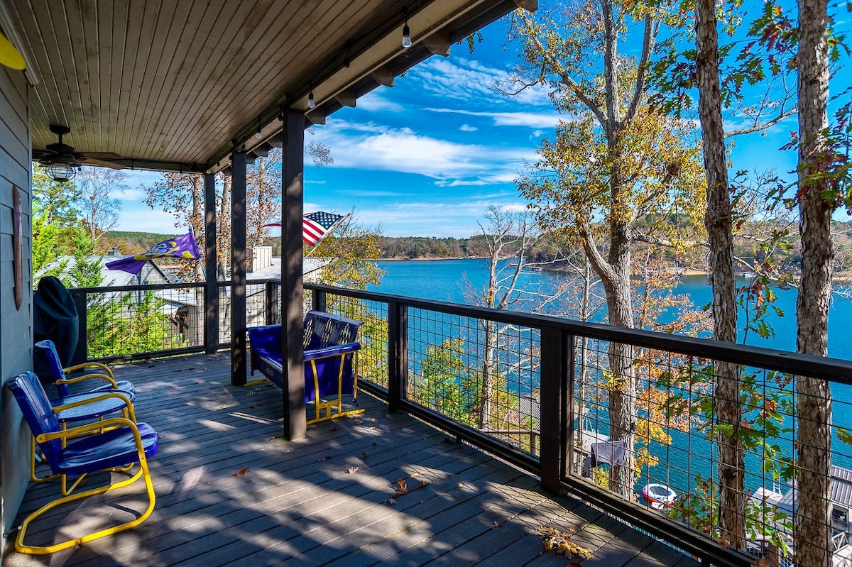 A covered deck with wooden flooring offers seating with blue chairs positioned against a railing. Trees surround the space, providing a view of the calm lake below, where the water reflects the clear blue sky and distant shore.