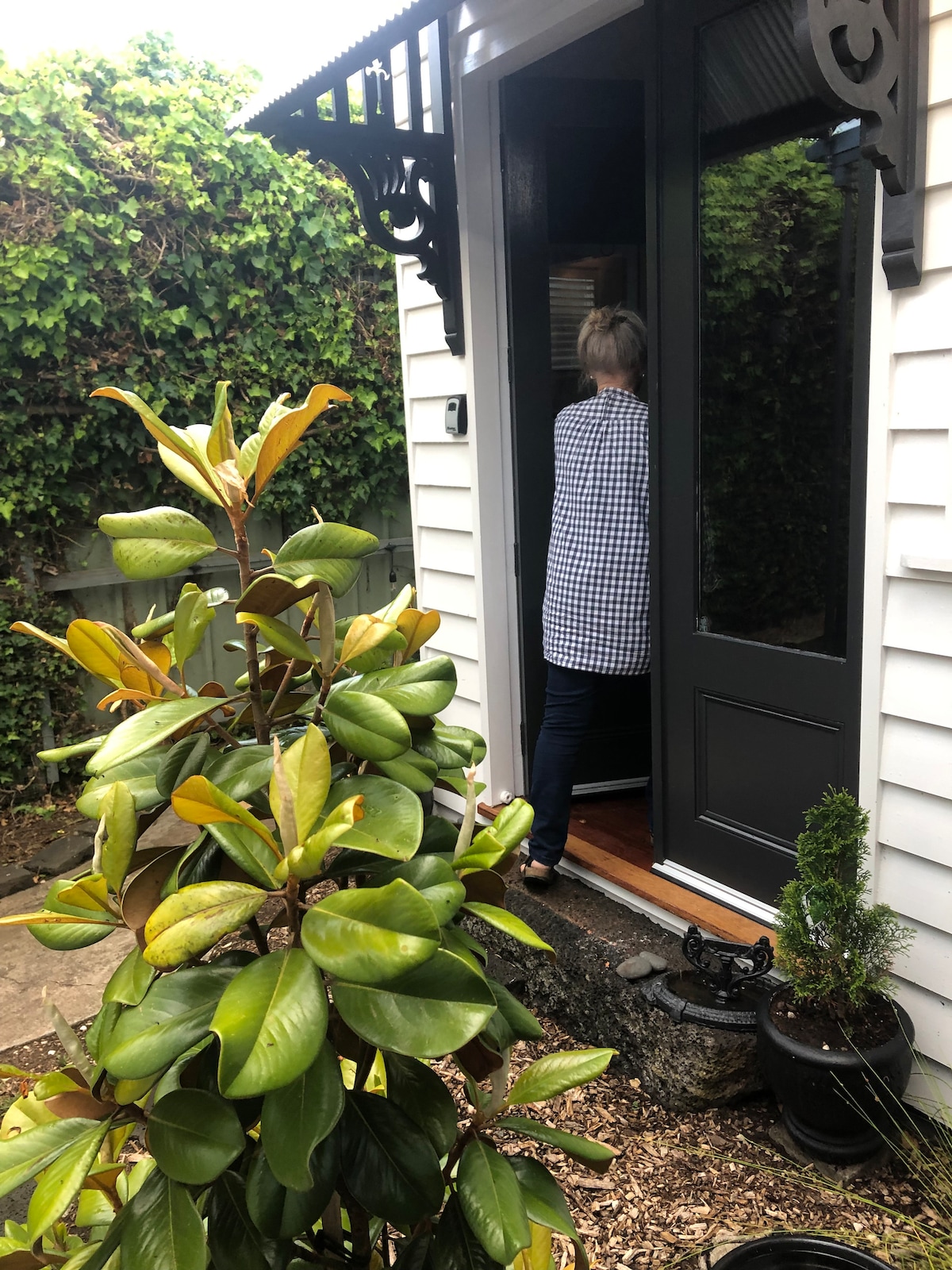 The entrance of the property is framed by a lush green bush and a potted plant. A person is seen entering through a large, dark door, surrounded by decorative architectural features and a wall of greenery.