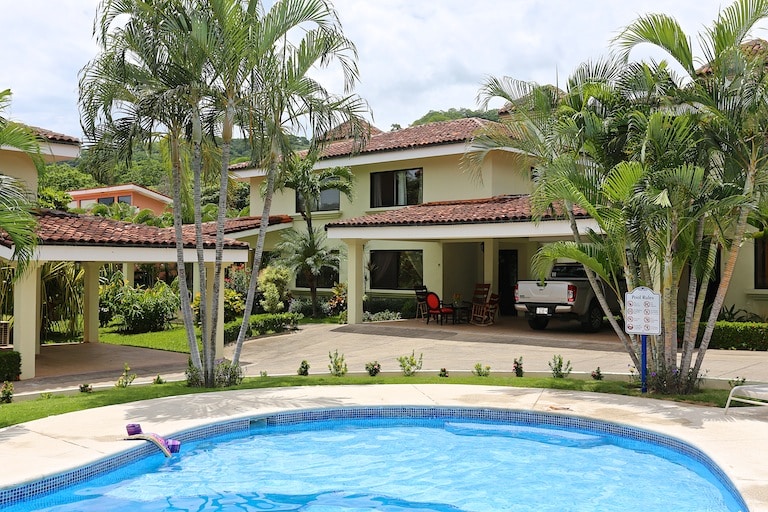 A clear view of the townhome complex showcases lush greenery and two inviting shared pools. The spacious outdoor area features palm trees, colorful landscaping, and the welcoming architecture of the two-story building, complementing the tranquil setting.
