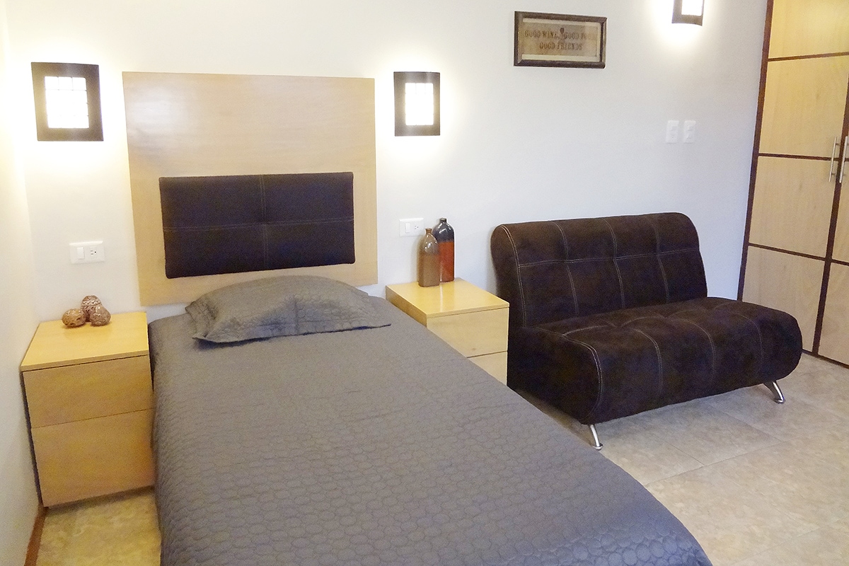 A compact bedroom features a single bed with a textured gray cover and a wooden headboard. Light fixtures are mounted on the wall, and a sleek black sofa bed is positioned against the side. Nightstands on either side provide additional storage.