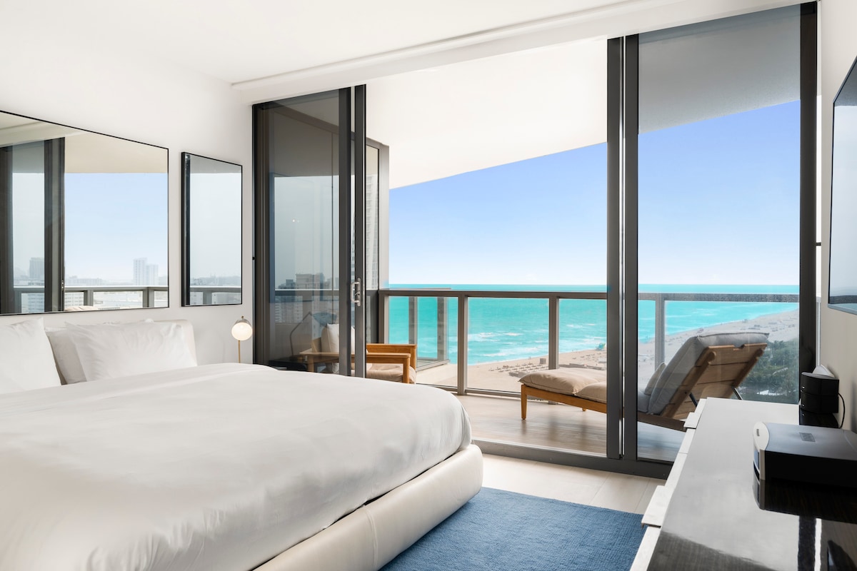 A well-appointed bedroom features a spacious bed and a flat-screen TV. Floor-to-ceiling glass doors open to a private balcony, revealing sweeping ocean views. A lounge chair sits on the balcony, offering a relaxing spot to enjoy the scenery.