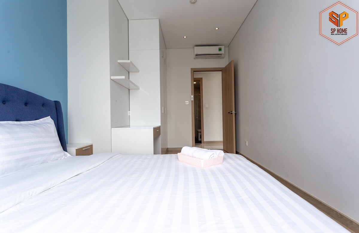 A bright sleeping area is featured with a spacious bed covered in white linens. A blue accent wall provides a contrast, while modern shelving and a simple wardrobe are positioned in the background. Soft natural light fills the room, creating a serene atmosphere.