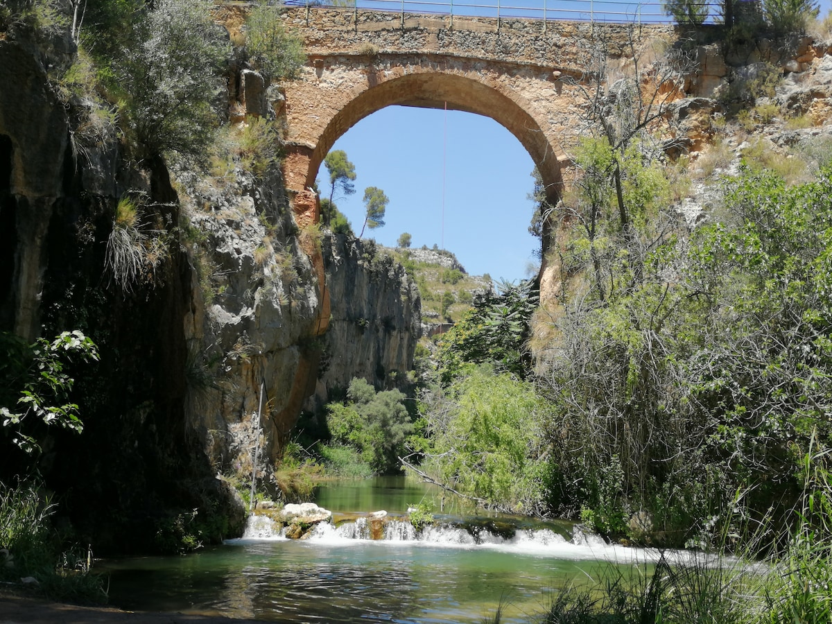 An arched stone bridge spans over a tranquil waterway, surrounded by lush greenery and rocky cliffs. Sunlight filters through the trees, casting gentle shadows on the water. The scene offers a serene depiction of the natural landscape, inviting exploration and appreciation of the environment.