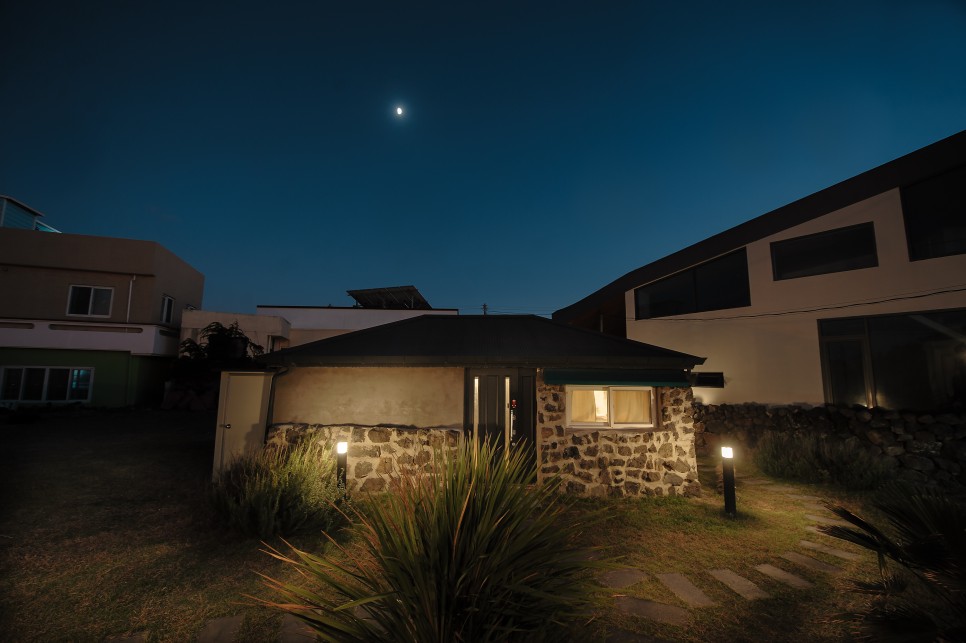 A stone house is illuminated softly at night, with its exterior showcasing the natural texture of the stone. The surrounding lush grass is complemented by pathway lights, creating a welcoming ambiance. The moon is visible above, adding a serene touch to the scene.