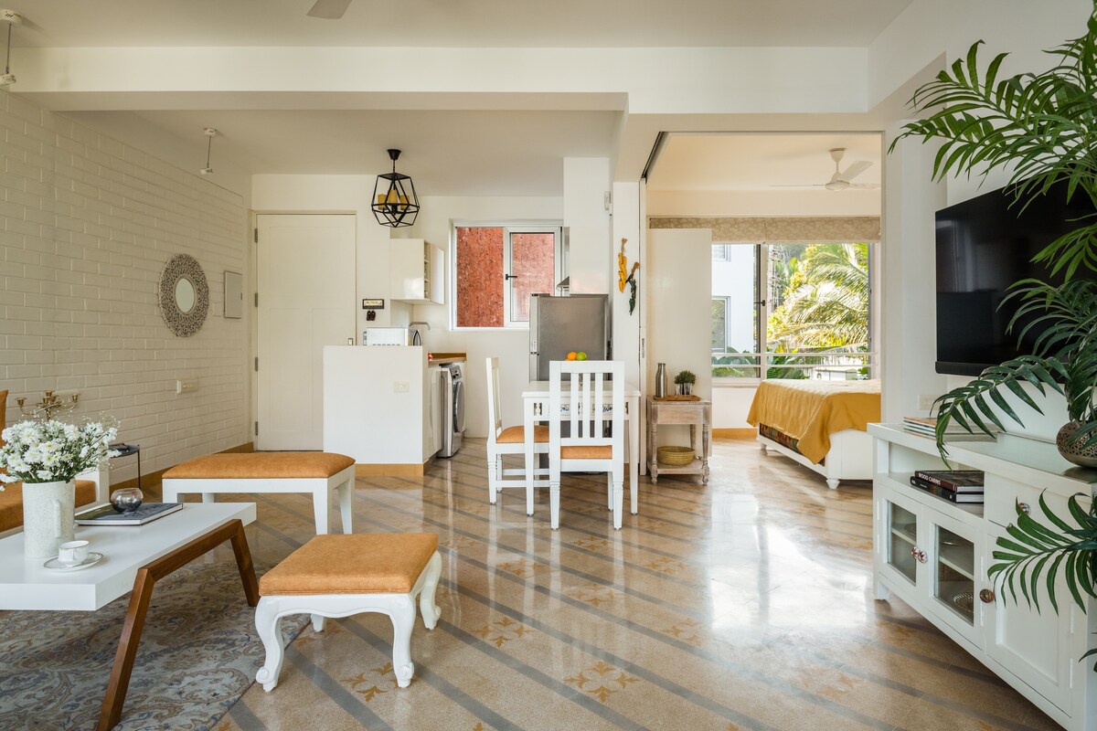 An open-plan living area is displayed, featuring a light-colored sofa and a coffee table. Dining chairs surround a white table. The kitchenette is equipped with modern appliances, and a glimpse of the balcony reveals greenery outside. Natural light brightens the space, creating an inviting atmosphere.