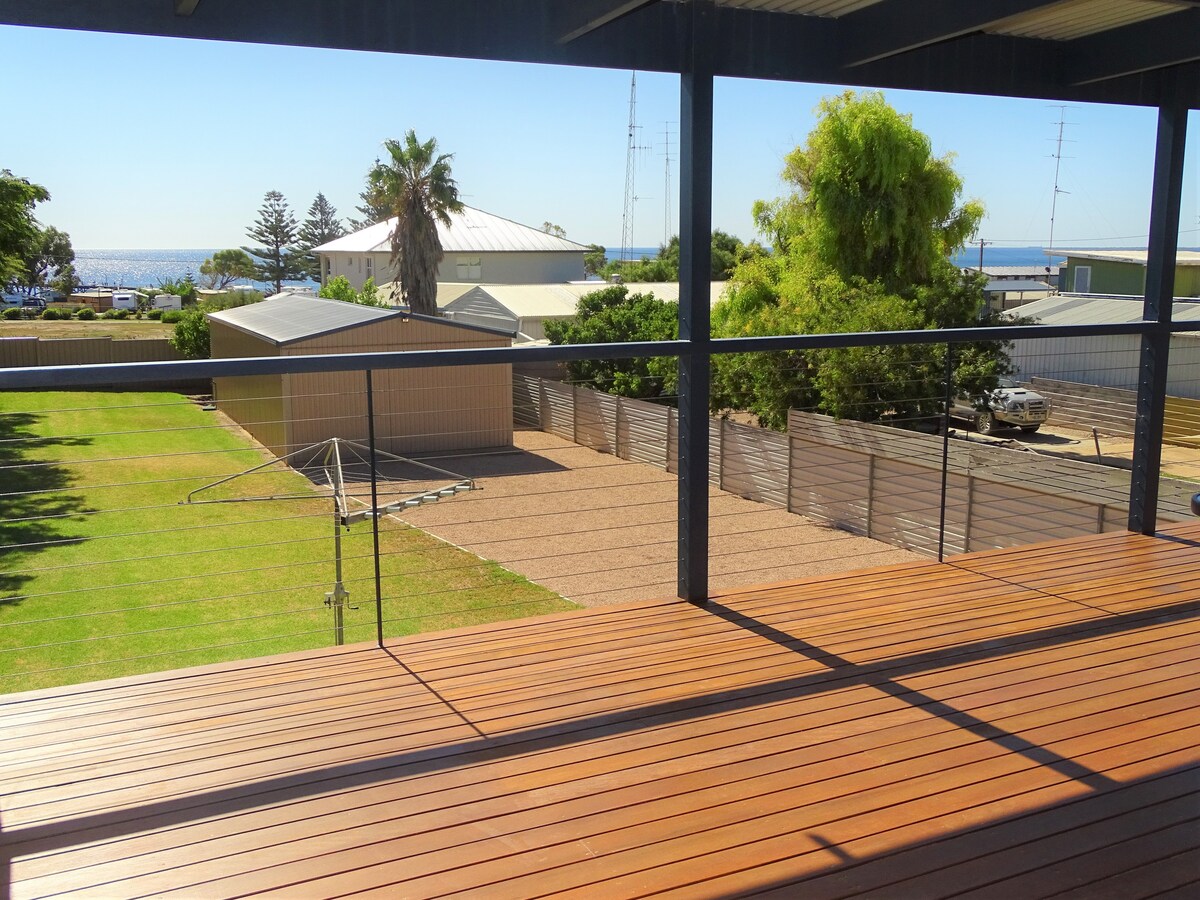 The upper balcony provides a view of the expansive fenced yard with a lawn area and outbuildings. Distant views of the coast can be seen, complemented by trees and a glimpse of the sea beyond.