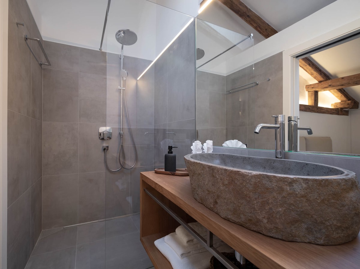 A modern bathroom features a walk-in shower enclosed with glass panels. A sculptural stone sink is positioned on a wooden vanity, complemented by neatly arranged towels below. Natural light is introduced through a skylight, highlighting the textured wall tiles.