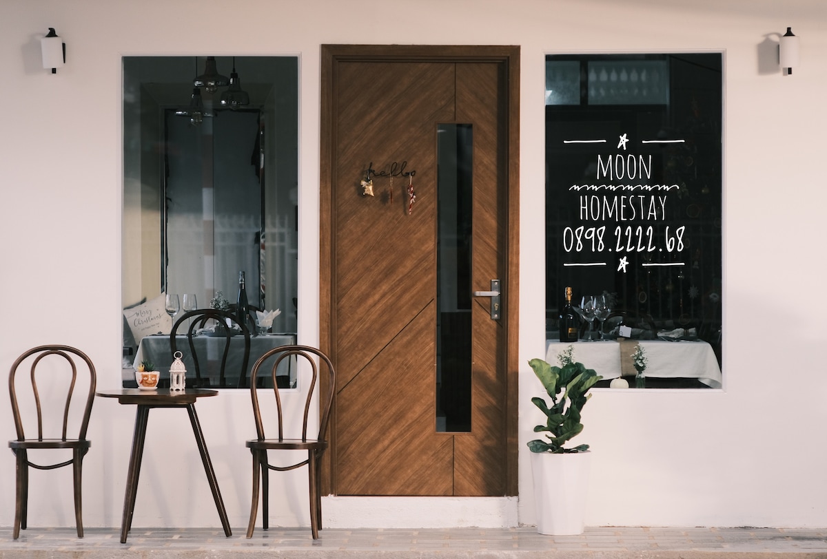 The entrance to Moon Homestay features a wooden door with glass panels. On either side of the door, a window displays the name and contact information. Two brown chairs sit around a small table, complemented by a potted plant near the entrance.