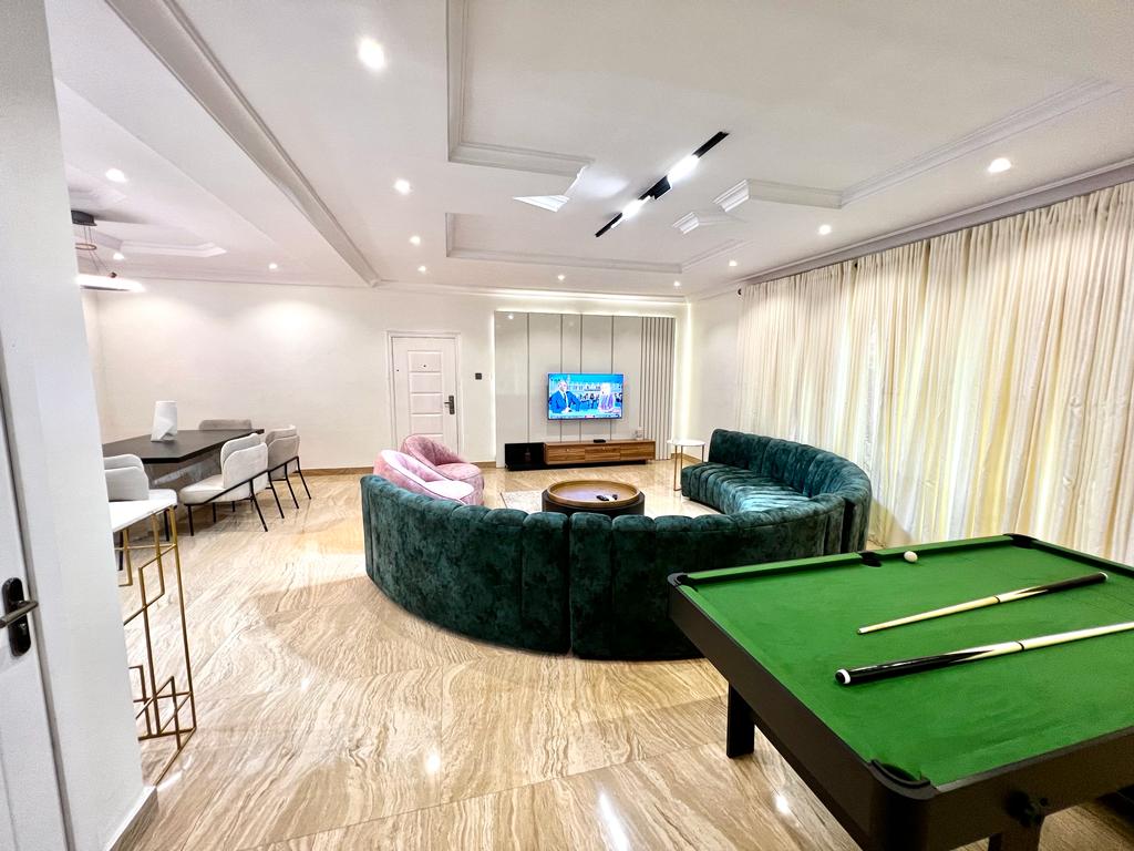 A spacious living area features a green sectional sofa and two cozy pink armchairs, arranged around a wooden coffee table. A large flat-screen TV is mounted on the wall, and a pool table is positioned nearby. Natural light filters through sheer curtains.