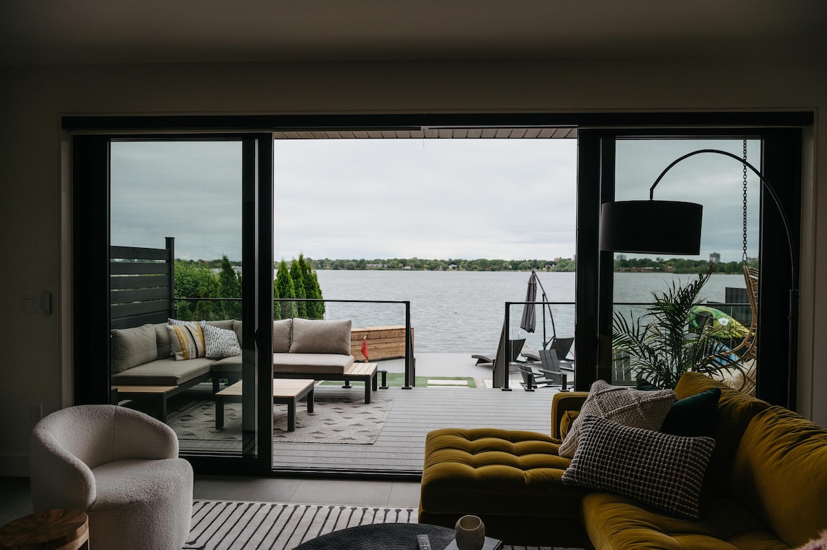 A modern living area features a large yellow sectional sofa and a cozy white accent chair. Expansive glass sliding doors provide a view of the waterfront and an outdoor seating area, blurring the lines between indoor and outdoor spaces.
