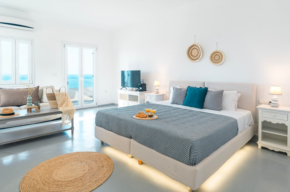 A spacious bedroom features a large bed with neutral bedding, flanked by bedside lamps. A soft area rug lies on the floor, and a seating area with a light-colored sofa is present. The room is brightly lit, showcasing views of the sea through the windows.