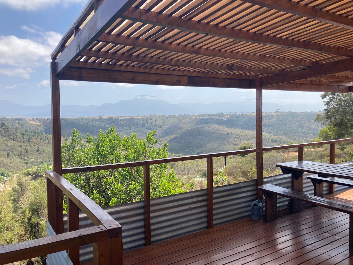 A spacious outdoor living area with wooden decking offers panoramic views of the surrounding hills and valleys. A table and benches are positioned on the deck, providing a comfortable space for relaxation. Lush greenery is visible in the foreground, enhancing the tranquil ambiance.