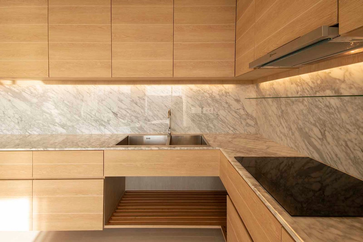 A modern kitchen is presented with wooden cabinetry and a smooth marble countertop. A stainless-steel sink is situated beneath a wall-mounted faucet. An electric cooktop is integrated into the counter, with under-cabinet lighting enhancing the workspace's brightness.