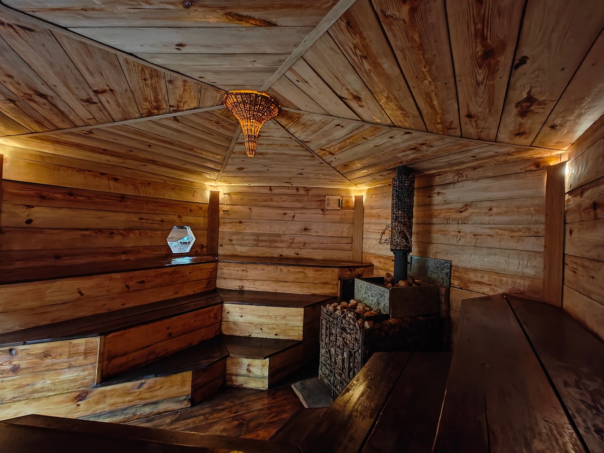 A cozy wooden sauna features warm wooden walls and benches, creating an inviting ambiance. Soft lighting enhances the natural materials, while a stone heater sits centrally, surrounded by smooth stones. An octagonal window allows for gentle light to filter in, contributing to the serene atmosphere.