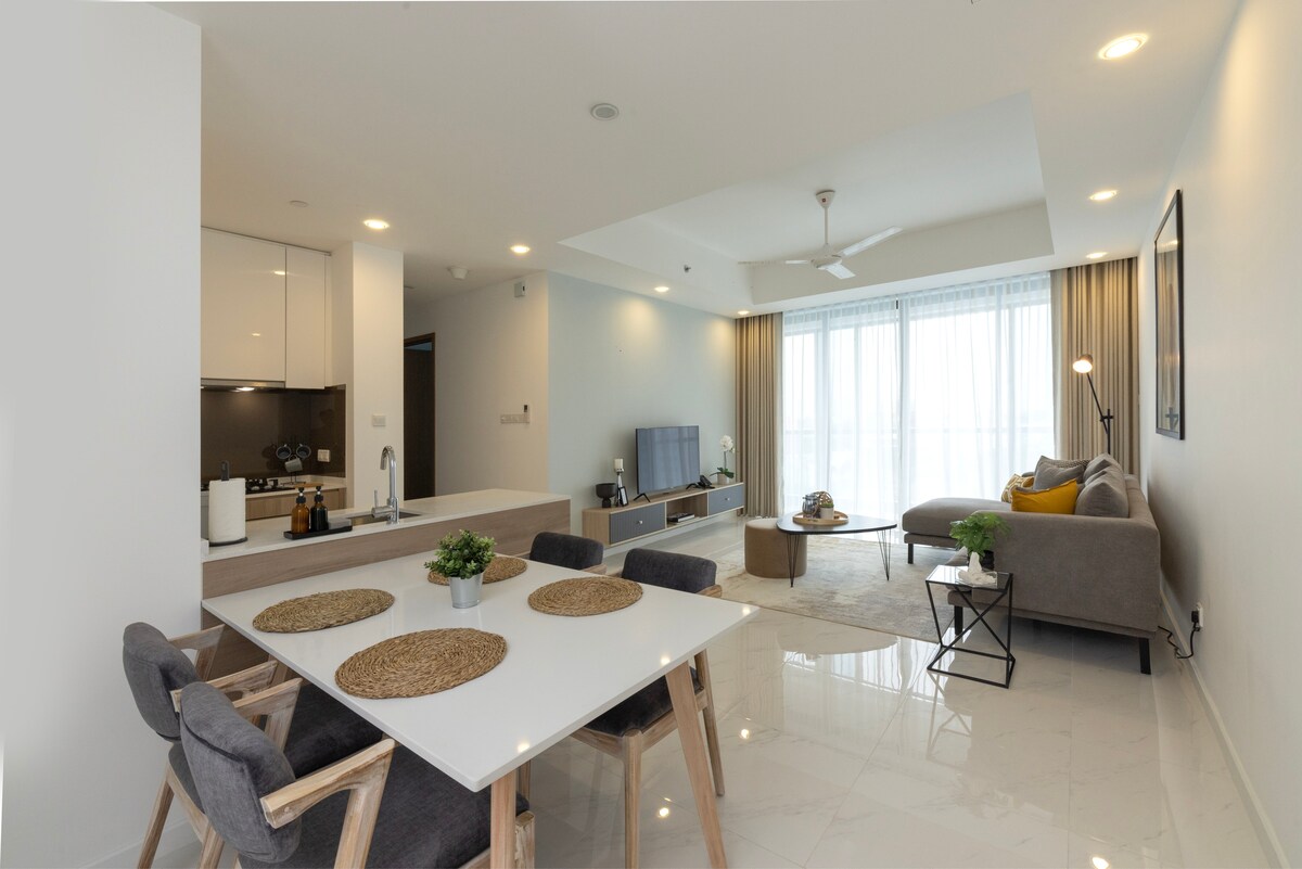 A modern living space is displayed, featuring a dining table set for four with woven placemats. A cozy seating area includes a gray sofa with bright cushions and a coffee table. Natural light fills the room through large windows, complemented by sleek, minimalist decor.