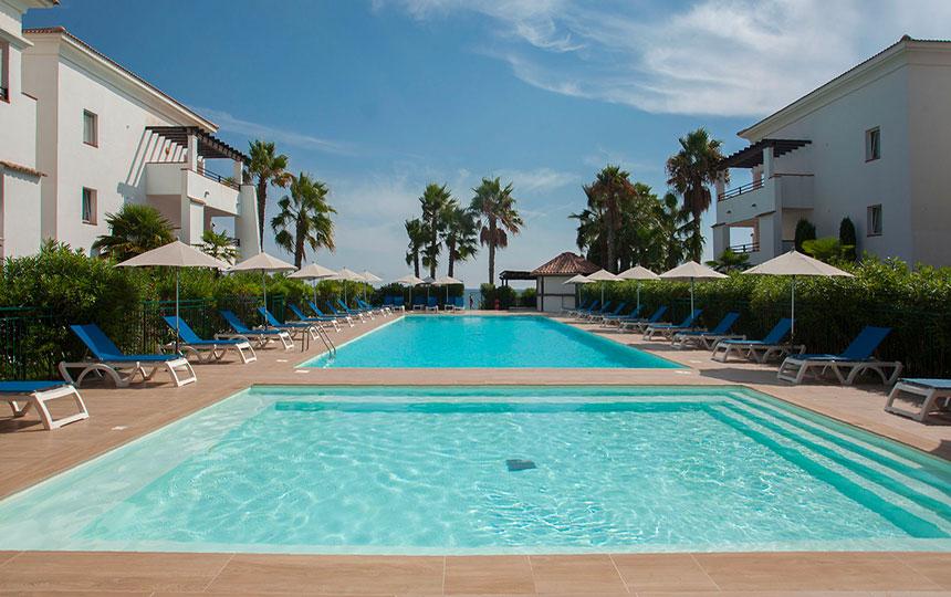 An outdoor pool area is showcased, featuring two connected swimming pools surrounded by lounge chairs and umbrellas. Lush palm trees line the perimeter, adding greenery to the tranquil setting. Bright blue skies and soft clouds provide a clear and inviting backdrop.