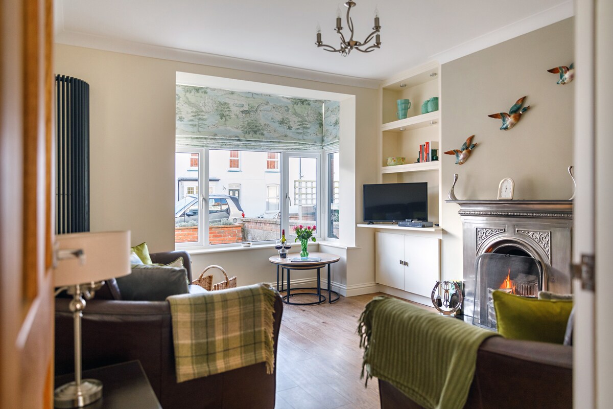 A sitting room features a square bay window letting in natural light. Two large leather sofas are arranged around a central coffee table. An original log-burning fireplace is highlighted, while a Smart TV is positioned on a wall unit. Decorative birds adorn the wall.