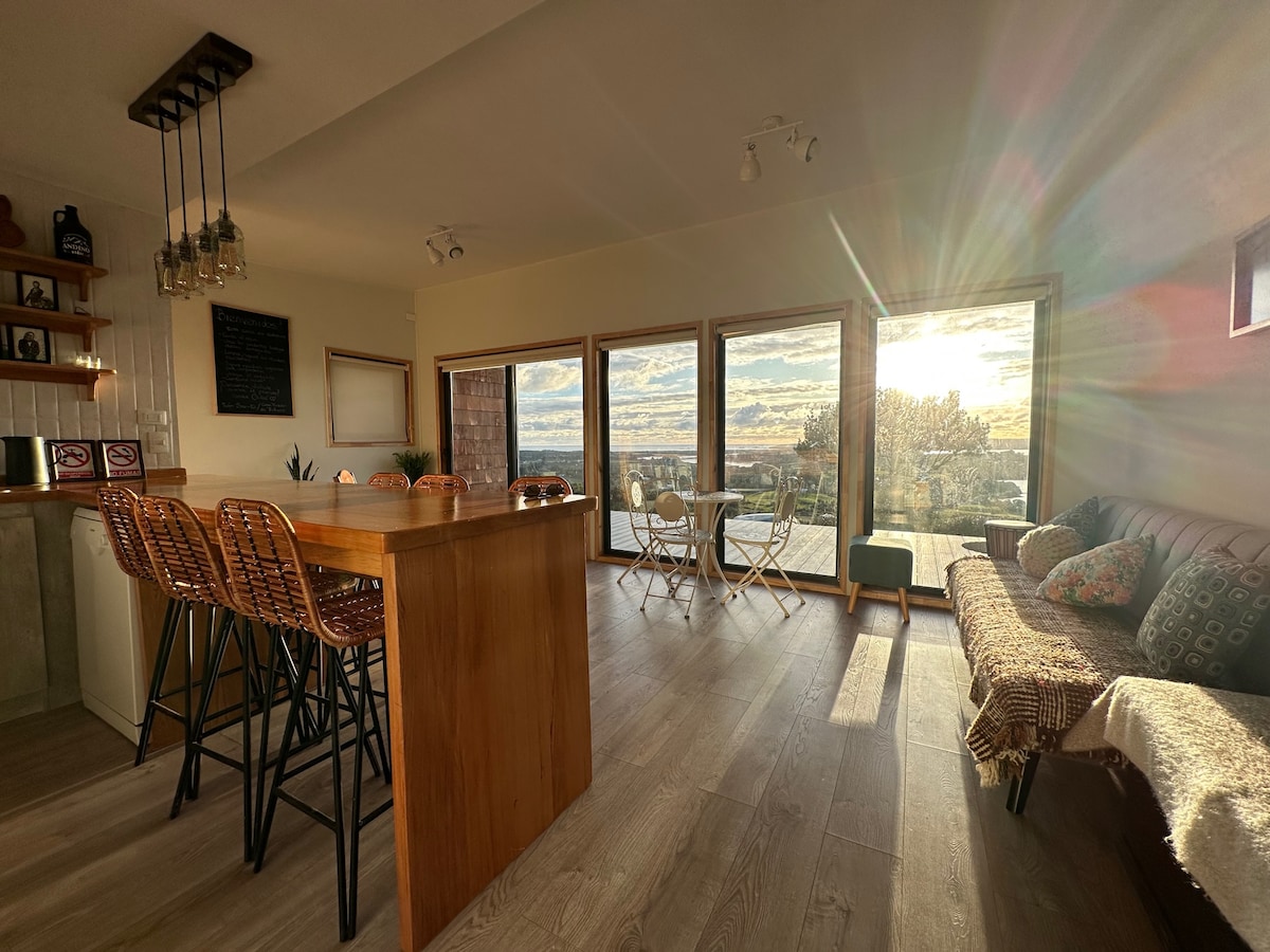 A bright, open-concept living area features large windows that offer scenic views of the surrounding landscape. A wooden bar with high stools provides a space for seating, while a cozy seating area showcases a woven blanket draped over a bench. Sunlight filters through, creating a warm ambiance.