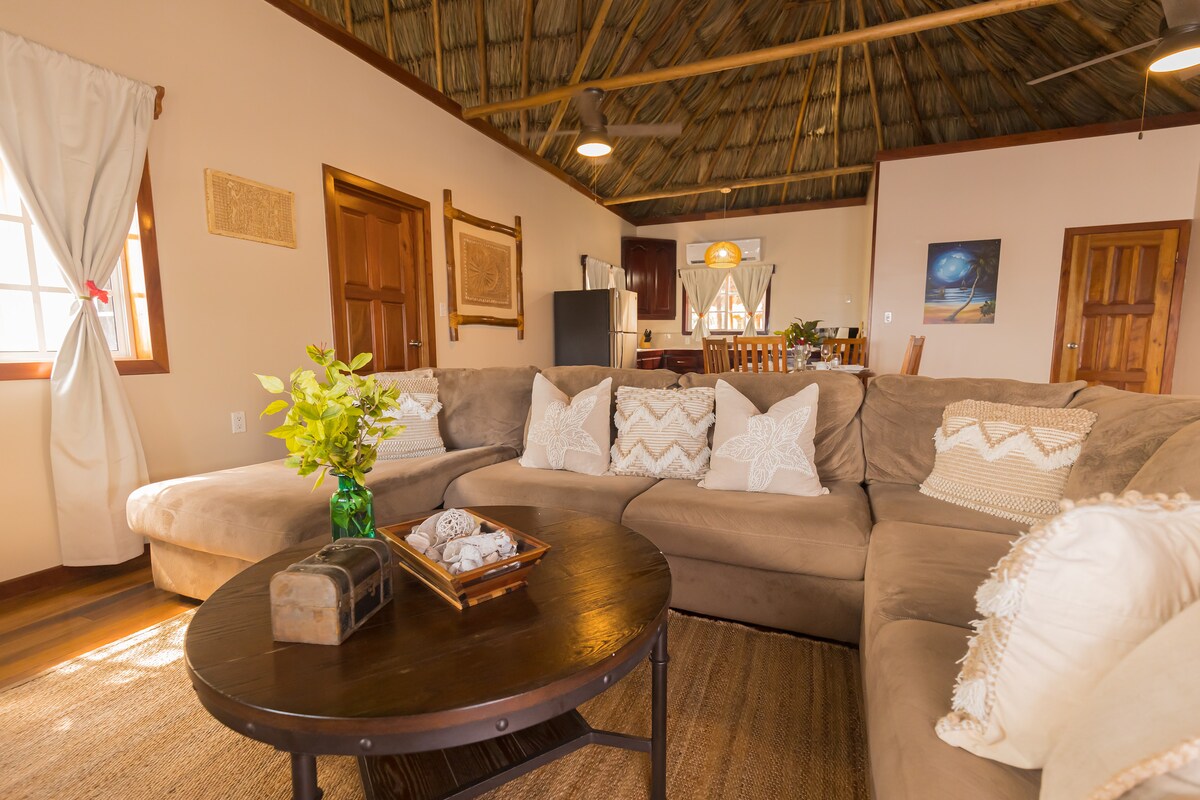 A spacious living area features an L-shaped sectional sofa adorned with textured pillows, inviting relaxation. A round coffee table holds decorative elements, while a ceiling with wooden beams enhances the open feel. Light streams through the windows, illuminating the warm tones of the furniture and decor.