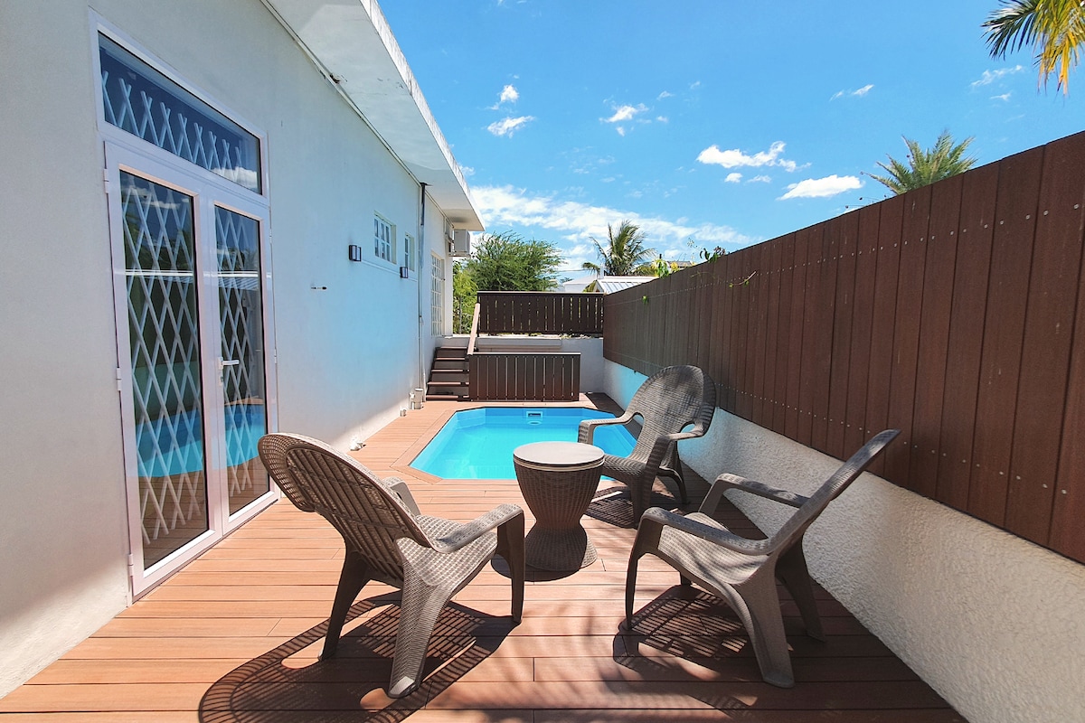 An outdoor seating area features four textured chairs arranged around a circular table, with a small private pool visible nearby. Sunlight illuminates the deck, and a wooden fence provides privacy while tropical plants are seen in the background.