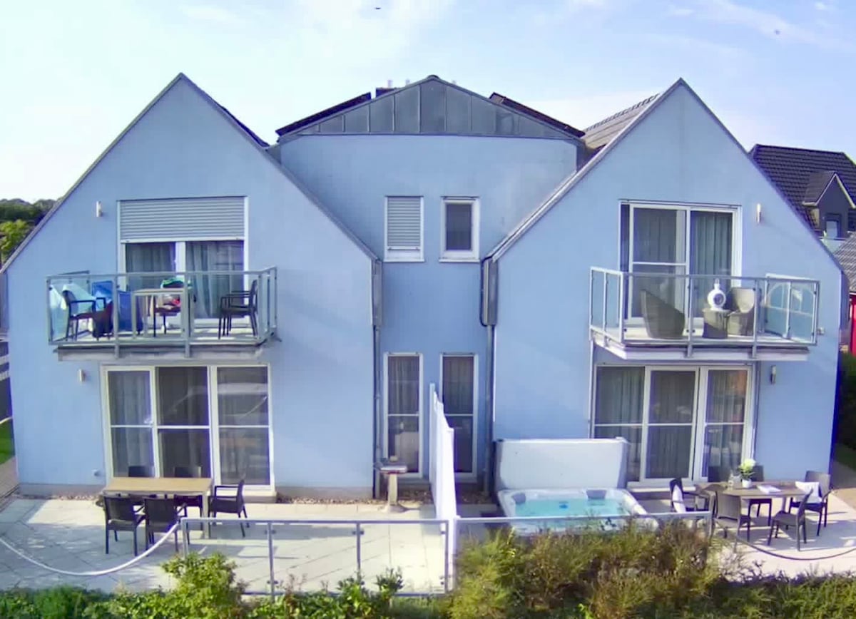 A light blue two-story building is presented, featuring two balconies with seating areas overlooking the garden. A shared outdoor patio area is visible, equipped with tables and chairs. A hot tub adds to the outdoor charm, surrounded by a manicured lawn.