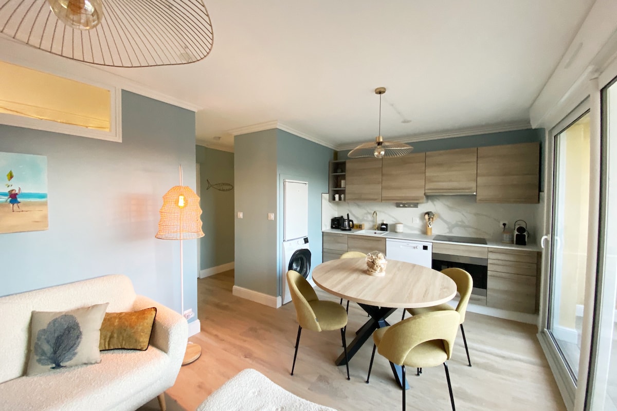 An open-plan living area features a modern kitchen with wooden cabinetry and a round dining table surrounded by yellow chairs. A light-colored sofa adds comfort, while a decorative lamp and artwork enhance the space. Large sliding doors provide natural light and outdoor access.