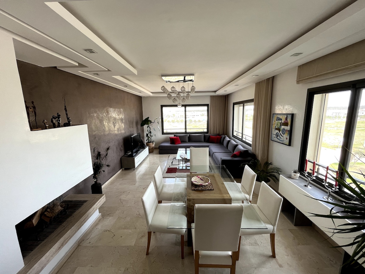 An elegantly arranged living space features a comfortable sectional sofa accompanied by red decorative pillows. A dining table with white chairs is set near large windows, allowing ample natural light. A modern fireplace is situated against a textured wall, contributing to the room's inviting ambiance.