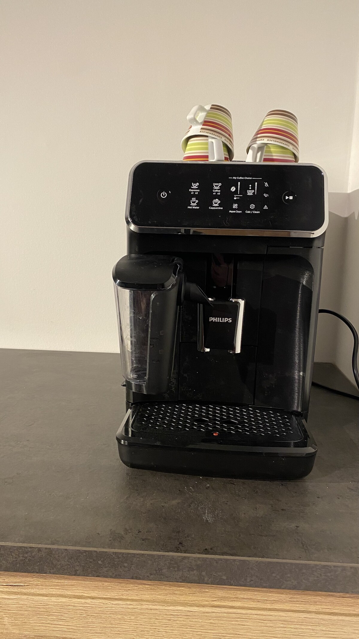 A modern coffee machine is positioned on a dark countertop, featuring a touch control panel and a transparent container for milk. Two colorful striped cups are placed on top of the machine, ready for use. The surrounding area is uncluttered, highlighting the appliance.