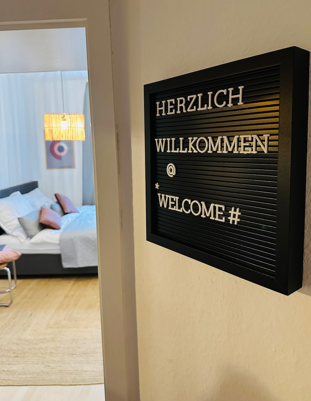 A welcome sign is displayed at the entrance, featuring a mix of German and English text. Soft lighting fills the space, revealing a cozy living area in the background, with minimalist decor and a gray sofa that invites relaxation.