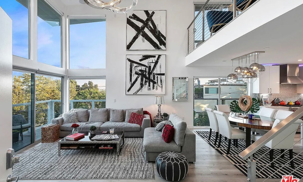 The modern living area features large windows allowing natural light to fill the space. A sectional sofa with decorative cushions is positioned alongside a low coffee table. Abstract artwork adorns the walls, while a dining table with multiple chairs is visible in the background.