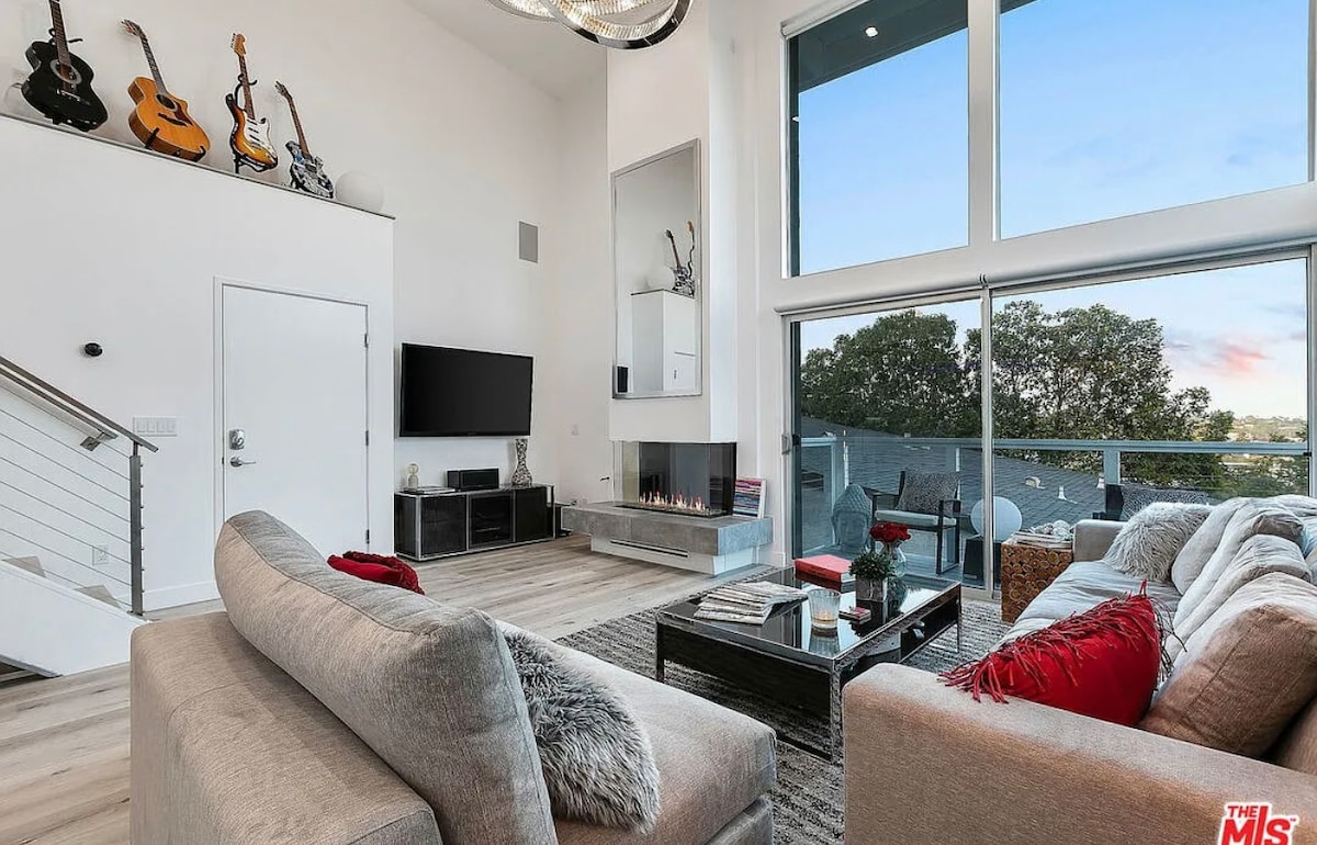The spacious living area features large windows that provide ample natural light and stunning views. A sleek television is mounted on the wall above a modern fireplace. Plush seating options include a large sectional sofa and accent chairs, complemented by stylish decor and a coffee table.