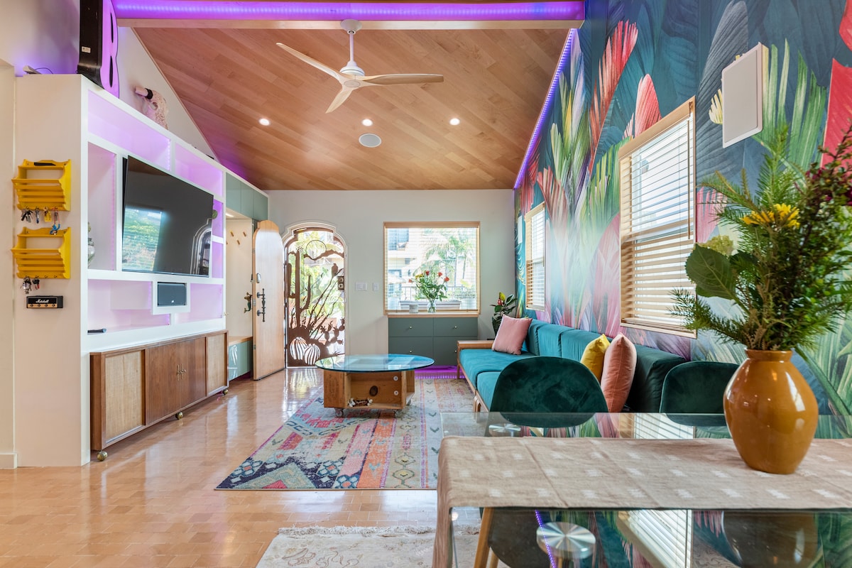 A vibrant living area displays a curved sofa in rich green tones, set against a tropical-themed wall. A round coffee table sits centrally on a colorful patterned rug. Natural light filters through large windows, enhancing the space's cheerful ambiance.