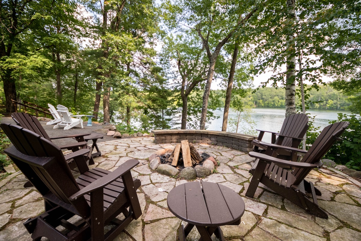An outdoor firepit area is surrounded by natural stone. Four dark wooden chairs are arranged around the firepit, accompanied by a small circular table. Lush greenery and a view of the lake provide a serene backdrop.